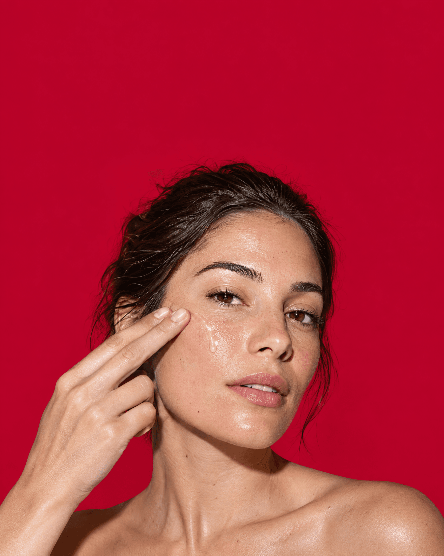 A woman smooths a clear gel-cream onto her cheek against a red backdrop.