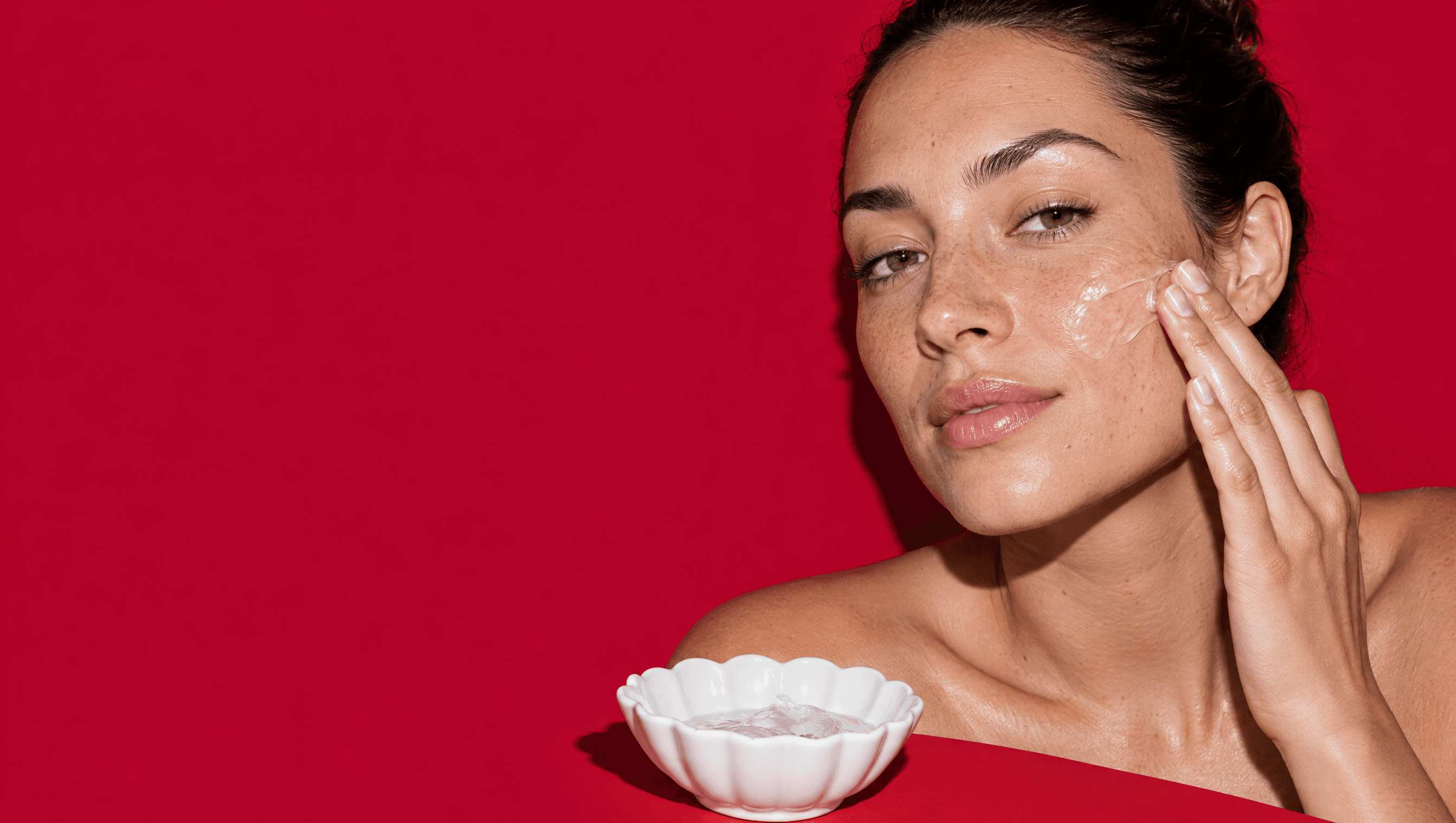 A woman smooths a clear gel-cream onto her cheek against a red backdrop.